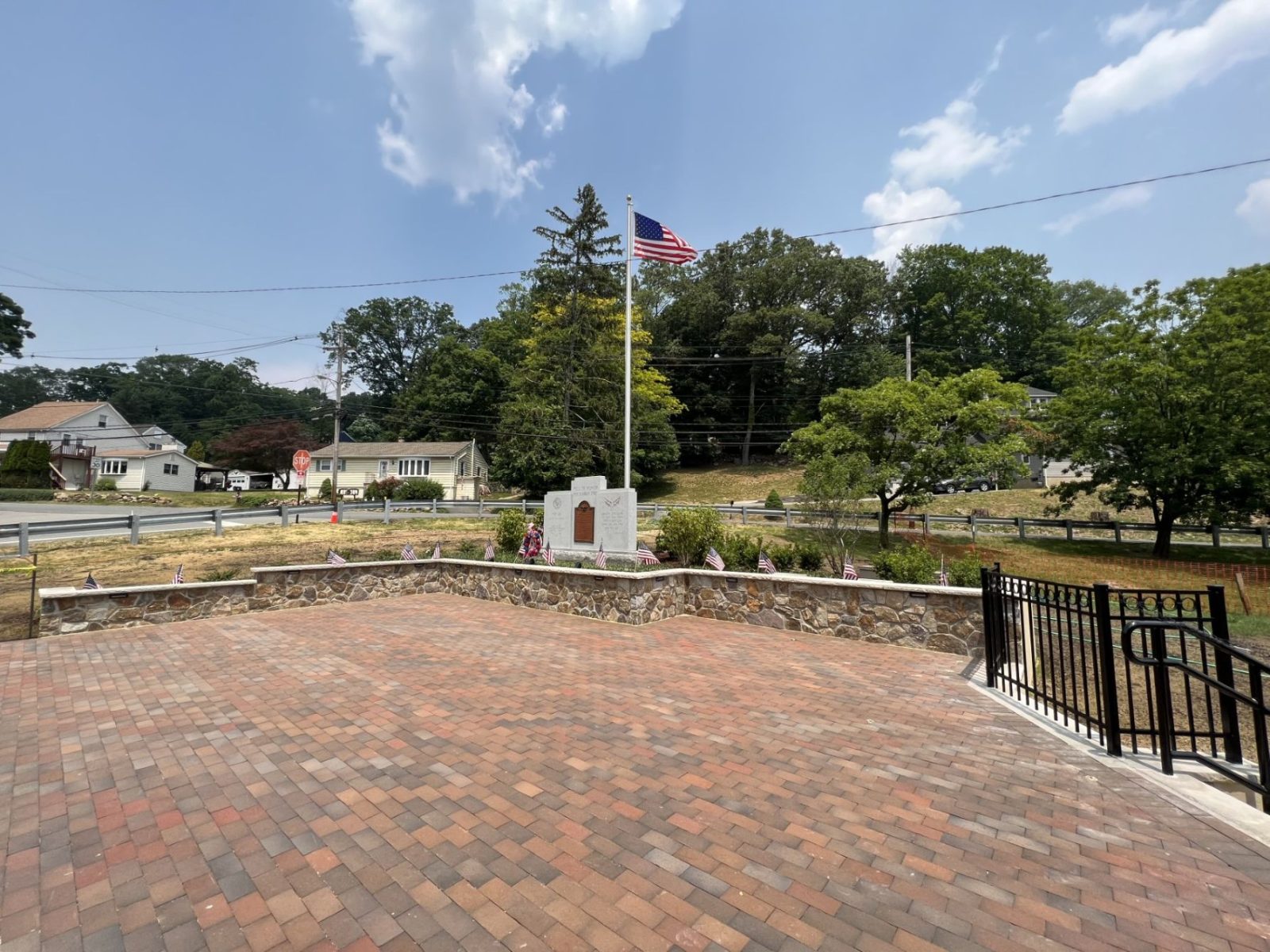 Park area with brick and flag pole surrounded by green trees.